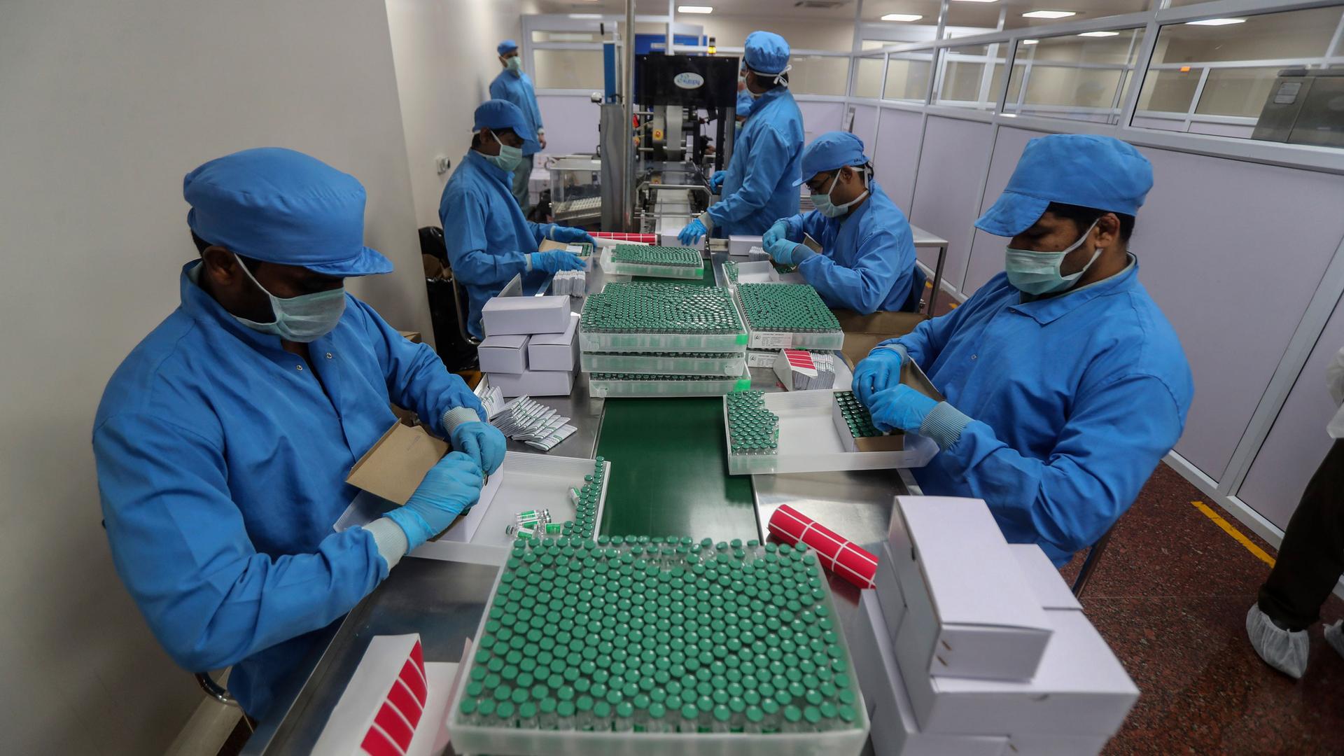 Several people are shown on either side of a long table wearing protective medical blue clothing and adding vials of COVID-19 vaccines to crates.