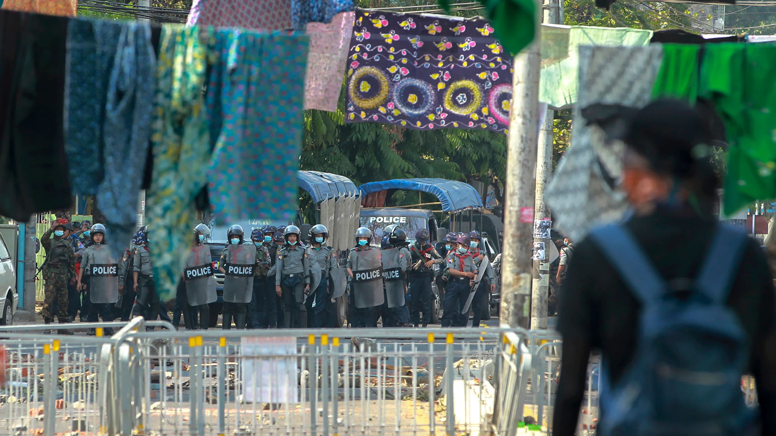 Security authorities are shown in the distance holding riot shield with several articles of clothing hanging on lines across the road.