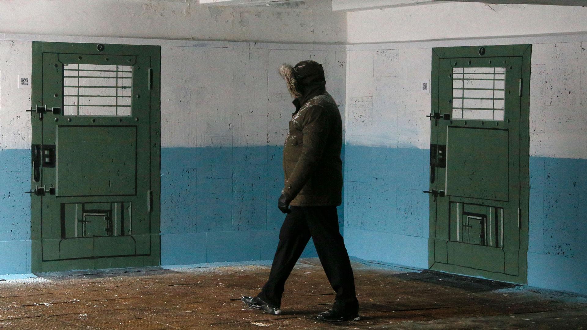 A person wearing a jacket with a hood up is shown walking past a wall with two prison doors painted on it.