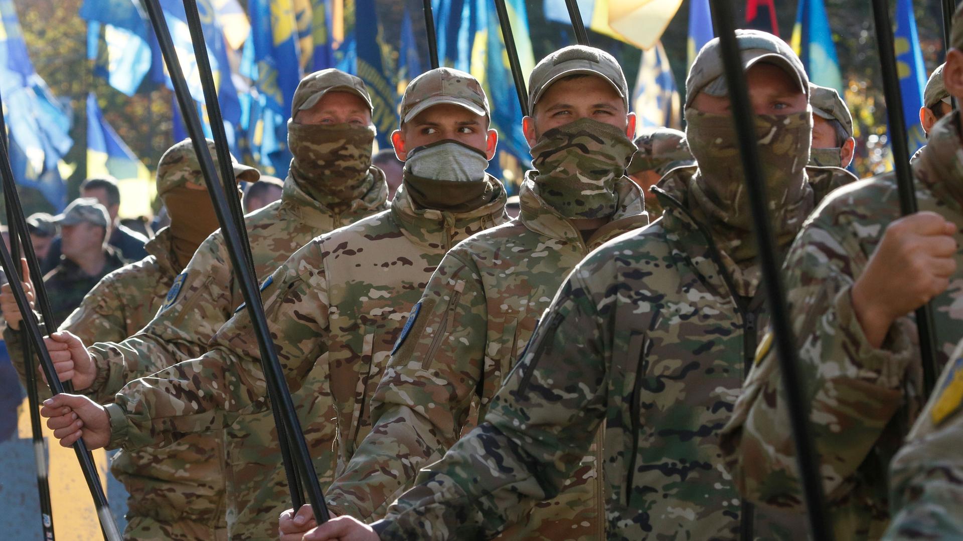 A line of men are shown wearing Ukrainian military fatigues and holding flag poles.