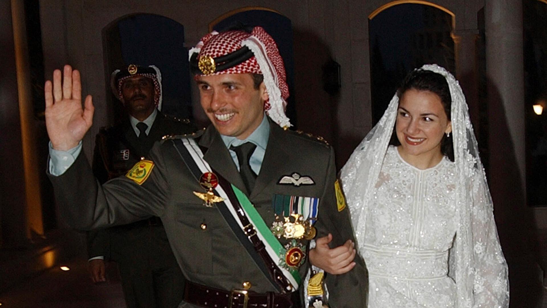 Prince Hamzah waves, wearing a military uniform walks with his bride, Prince Noor, wearing a white wedding dress and they both smile. 