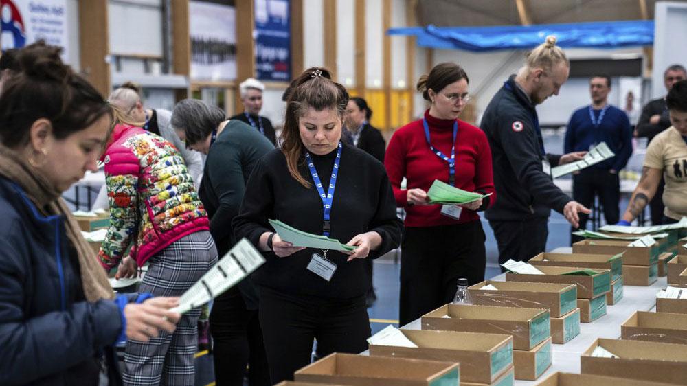 A small crowd of people are shown examining green ballots next to rows of boxes.