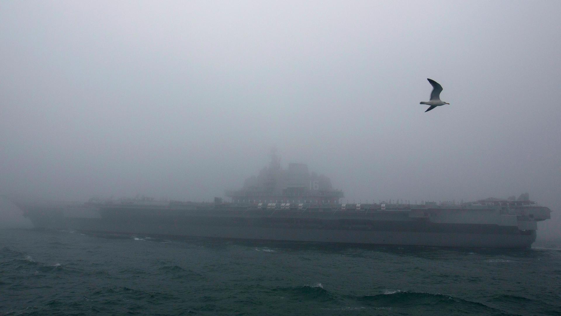 A large military vessel is shown in the distnace amid foggy weather conditions.