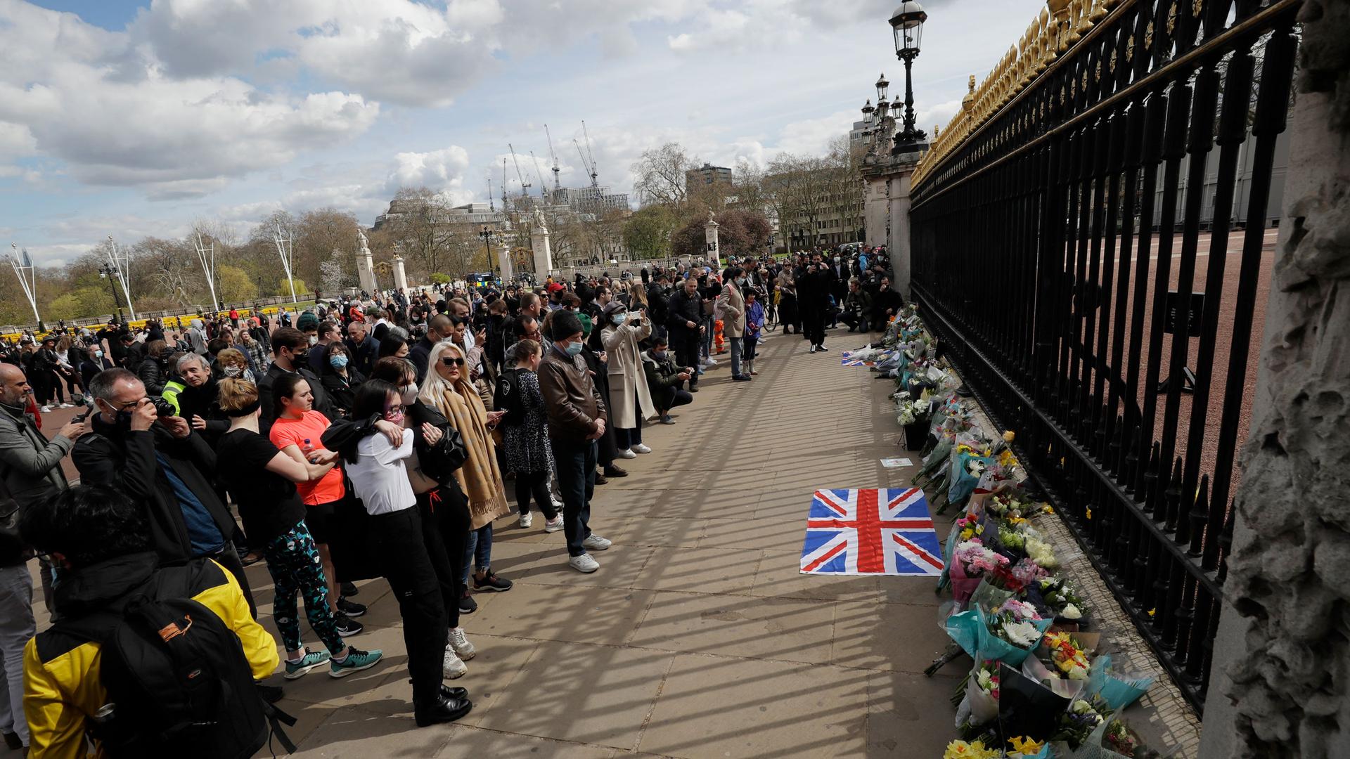 A large crowd of people are shown outside of the tall black iron gates of Buckingham Palace.