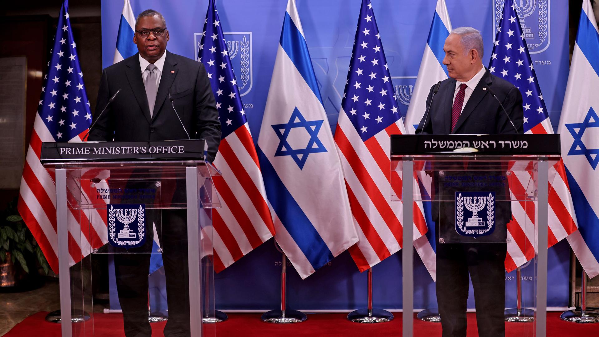 US Defense Secretary Lloyd Austin and Israeli Prime Minister Benjamin Netanyahu are shown each standing behind podiums with US and Israeli flags in the background.