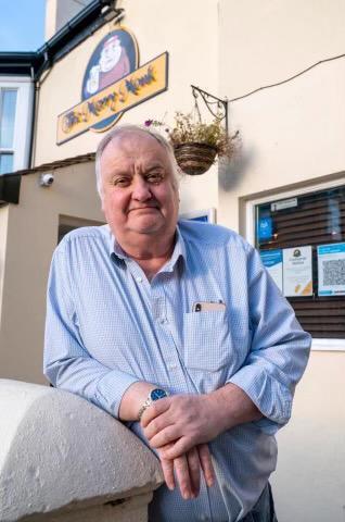 A rotund white man with gray hair smiles and poses for a photo wearing a blue shirt outside one of his pubs, The Merry Monk.
