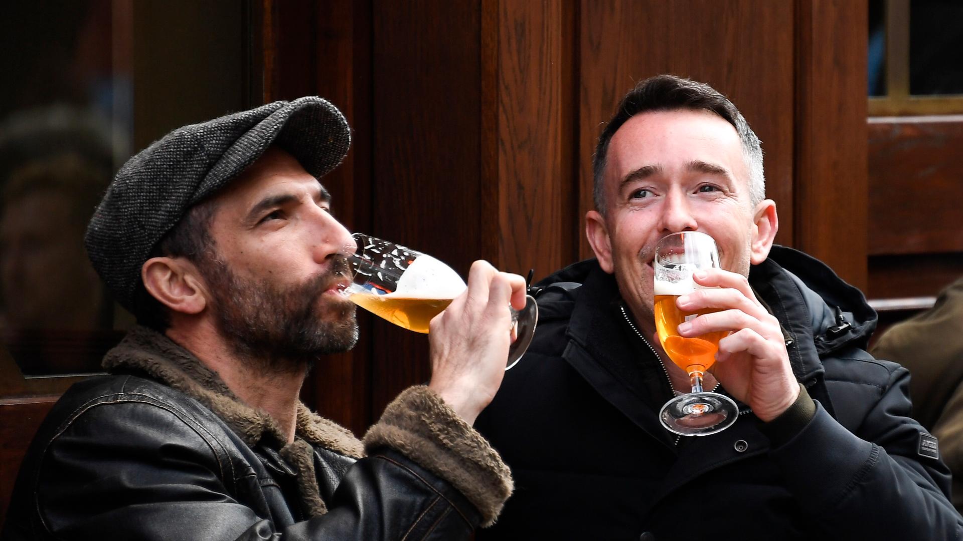 Two white men wearing dark jackets drink beer outdoors at a pub in London. 