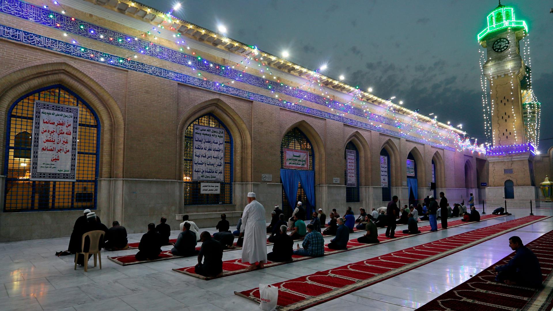 Several rows of people are shown kneeling next to a building with bright lights strung across the top.