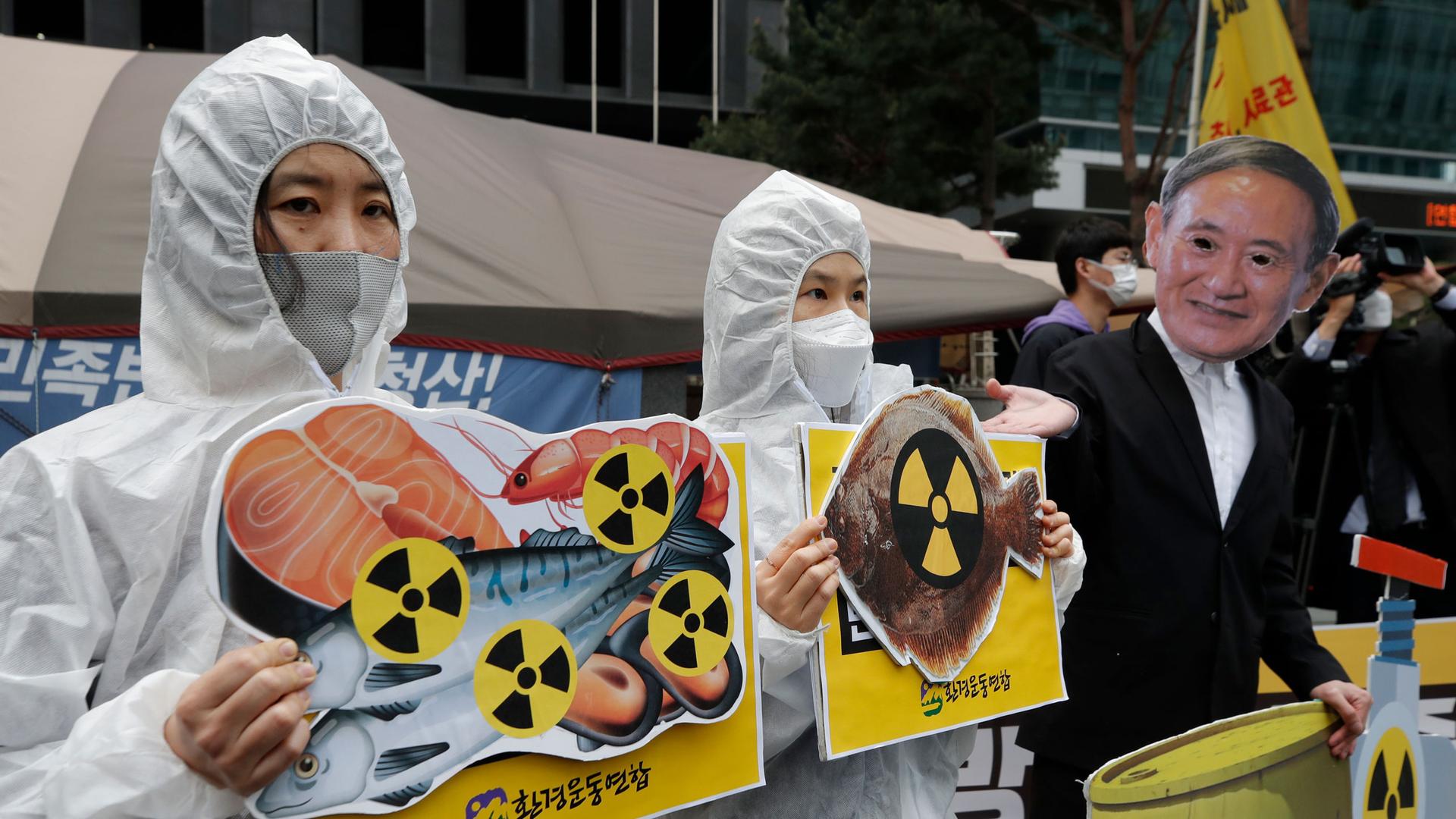 A protester is shown wearing a white radiation protection suit and holding a sign with cuts of fish and the radioactive symbol.