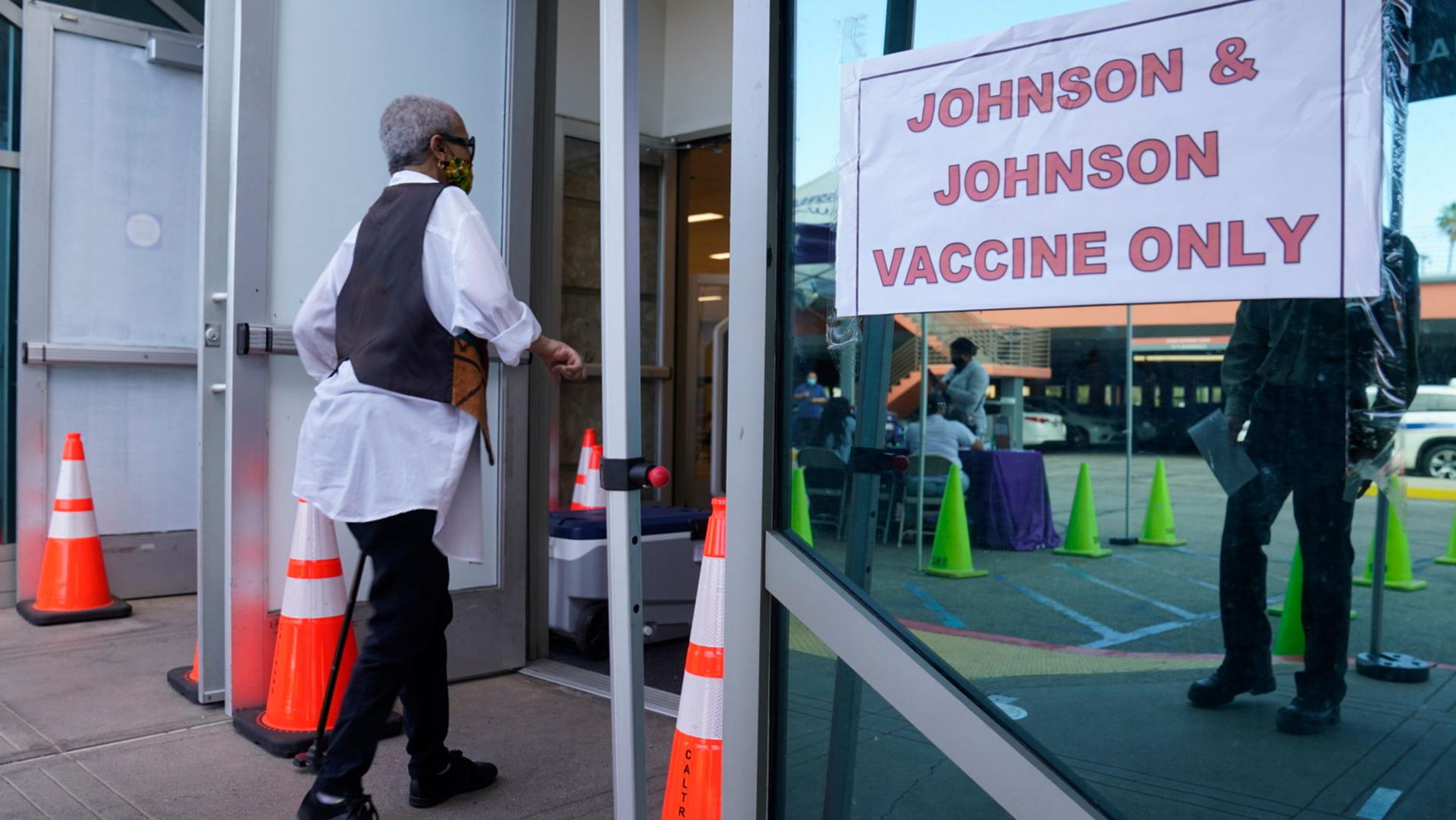A person is shown wearing a dark vest and white shirt and walking with a cane into a facility with a Johnson & Johnson sign on the outside.