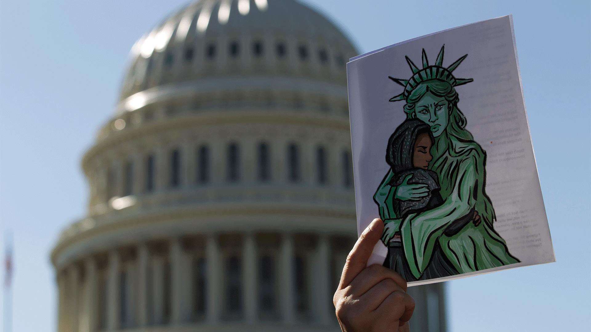 A hand holds up a cartoon in front of the Capitol dome of the Statue of Liberty hugging a Muslim hijabi girl.