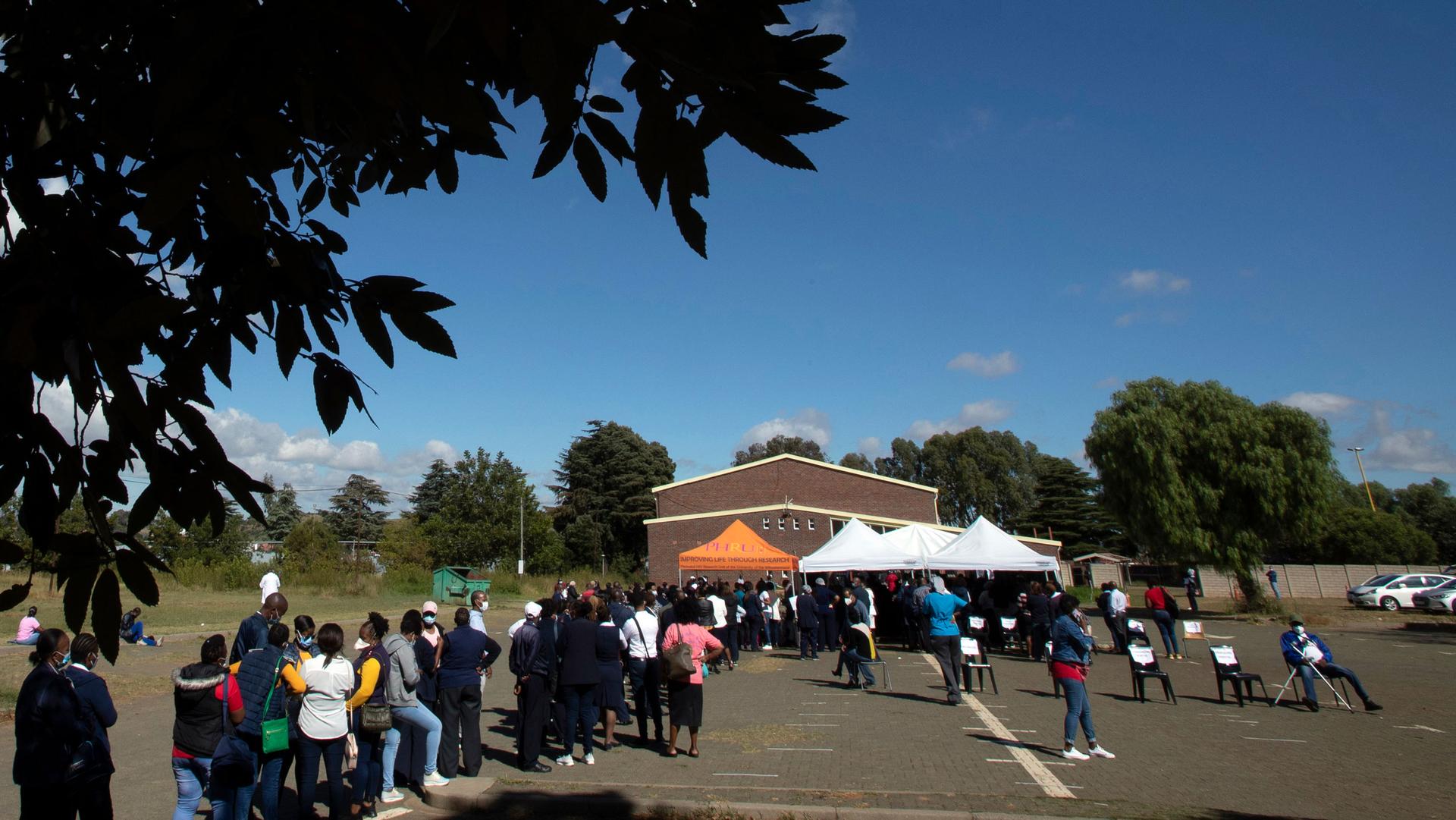 A long line of people are shown outside in a pkarking lot with an orange tent in the distance.
