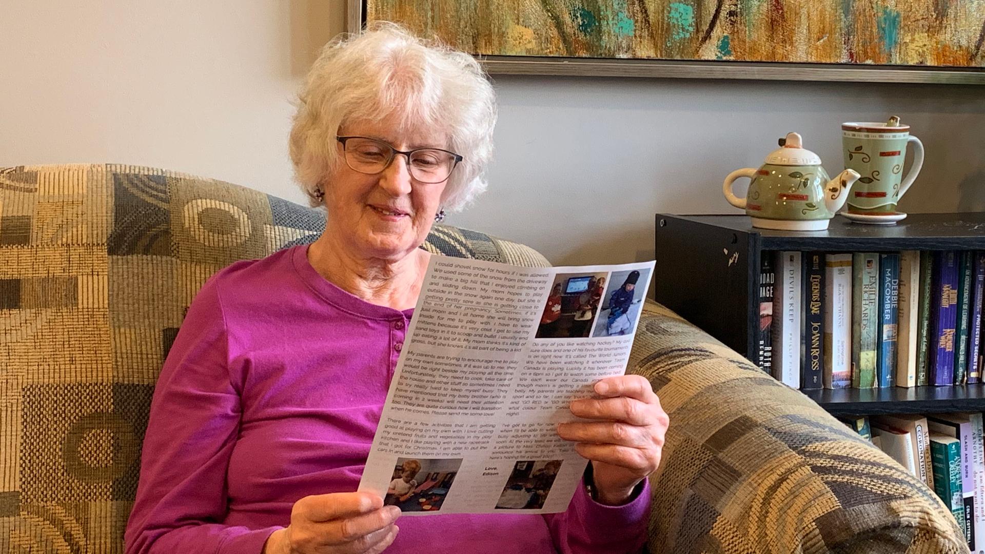 A 91-year-old white woman with gray hair, wearing glasses and a pink top, sits on a sofa and reads a newsletter. 