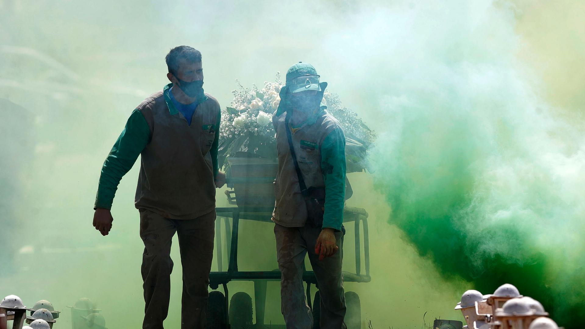 Two cemetery workers are shown pulling a casket on wheels with ceremonial green smoke surrounding them.