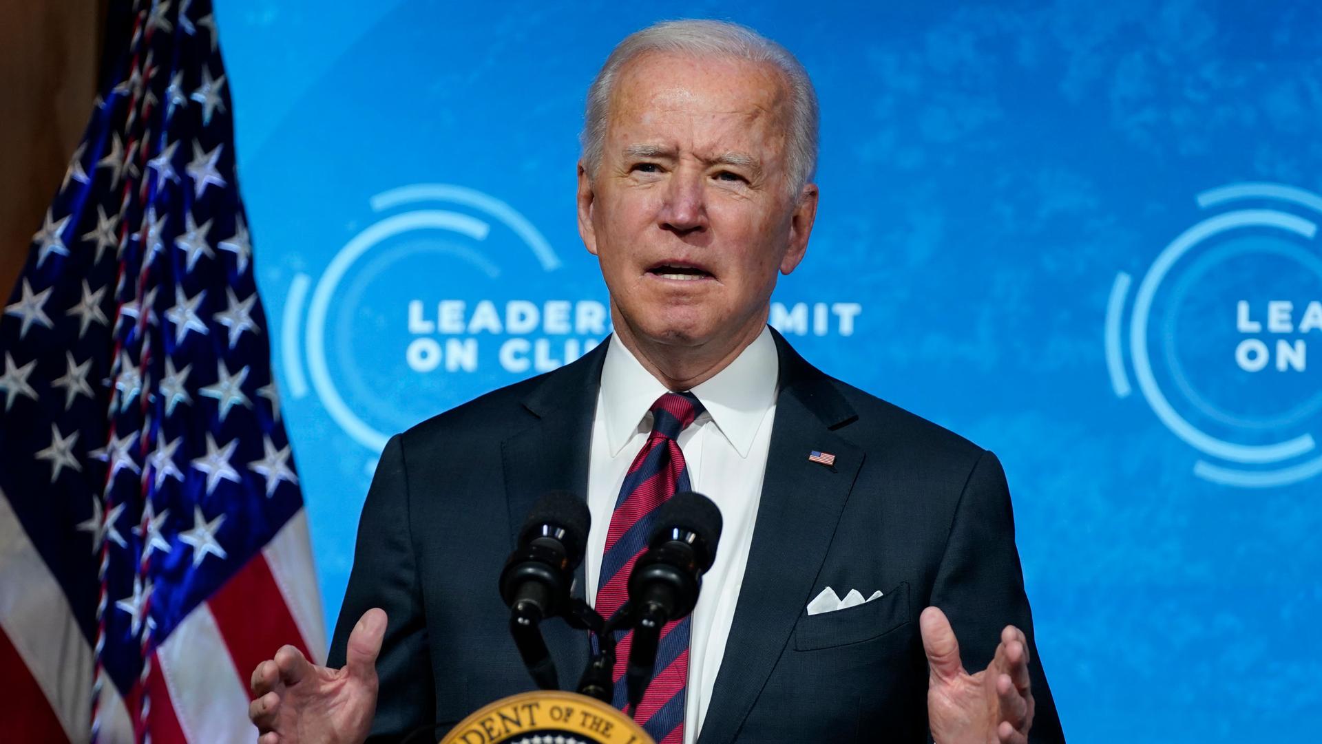 US President Joe Biden is shown standing at a podium wearing a suit and tie with an US flag to his right.