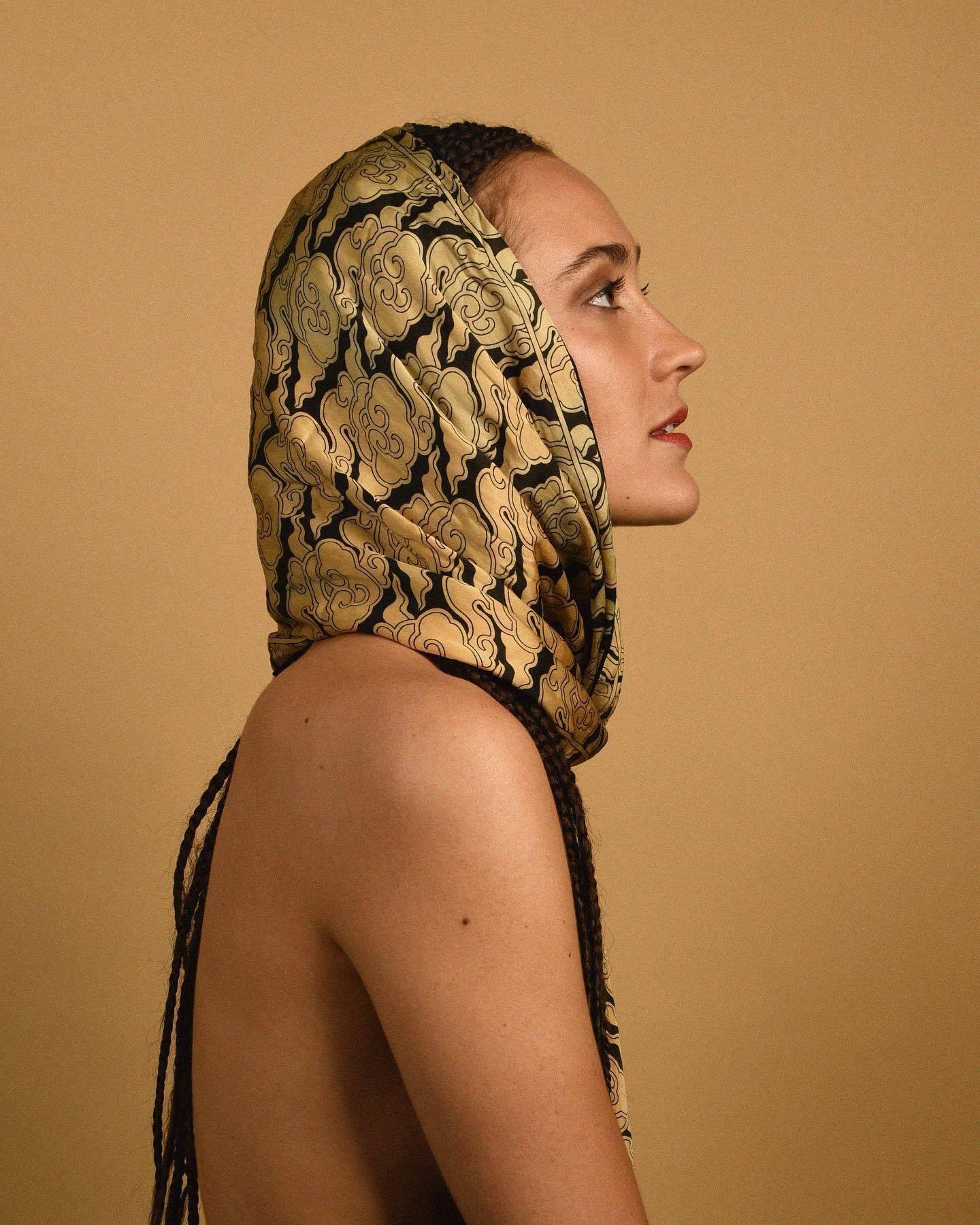Justine Mauvin, also known as Sibu ManaÏ, featured in a profile pose wearing a gold and black headscarf with light-skinned shoulders showing from the side.