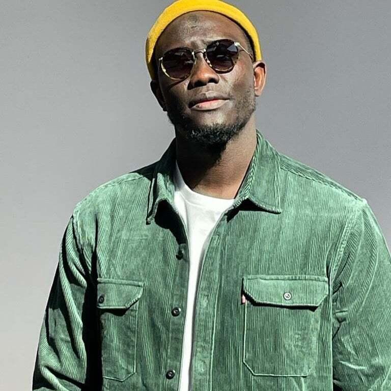 Senegalese artist Lass, a Black man wearing a green jacket, white shirt underneath, with a yellow hat and sunglasses, poses for a photo.
