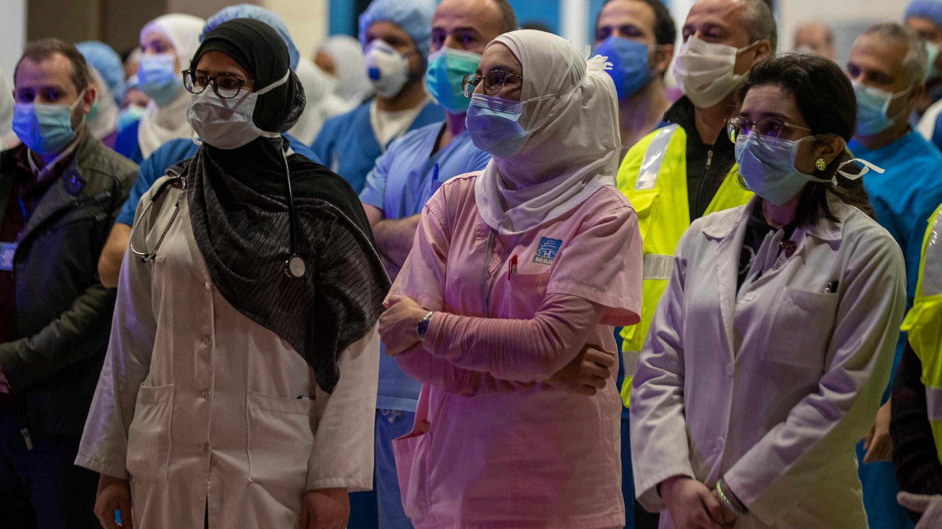 Three women health care workers in uniform, wearing headscarves and masks, stand and listen among a crowd of medical workers. 