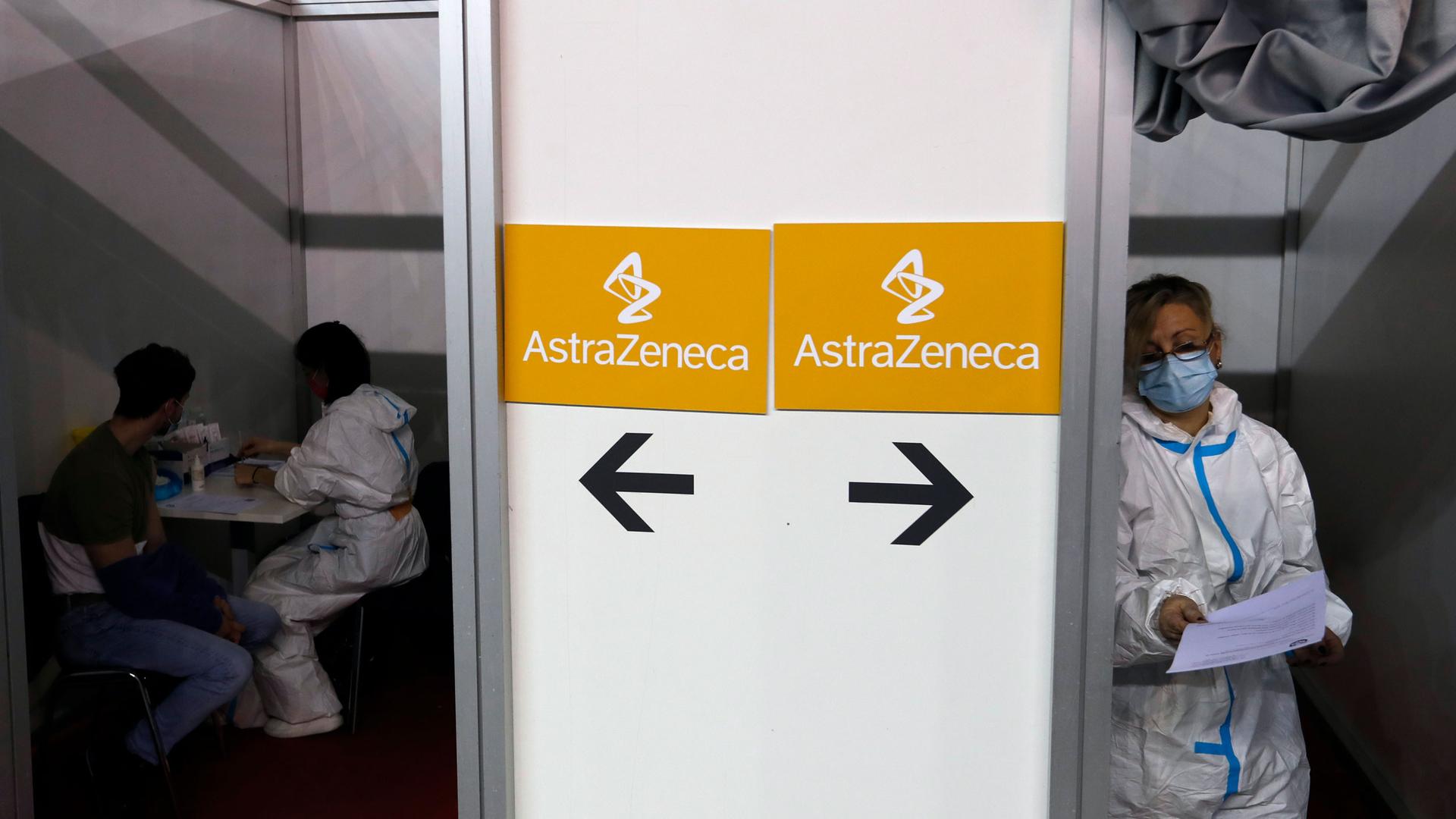 Medical professionals are shown on either side of a wall that has signs for the AstraZeneca vaccine posted on it.