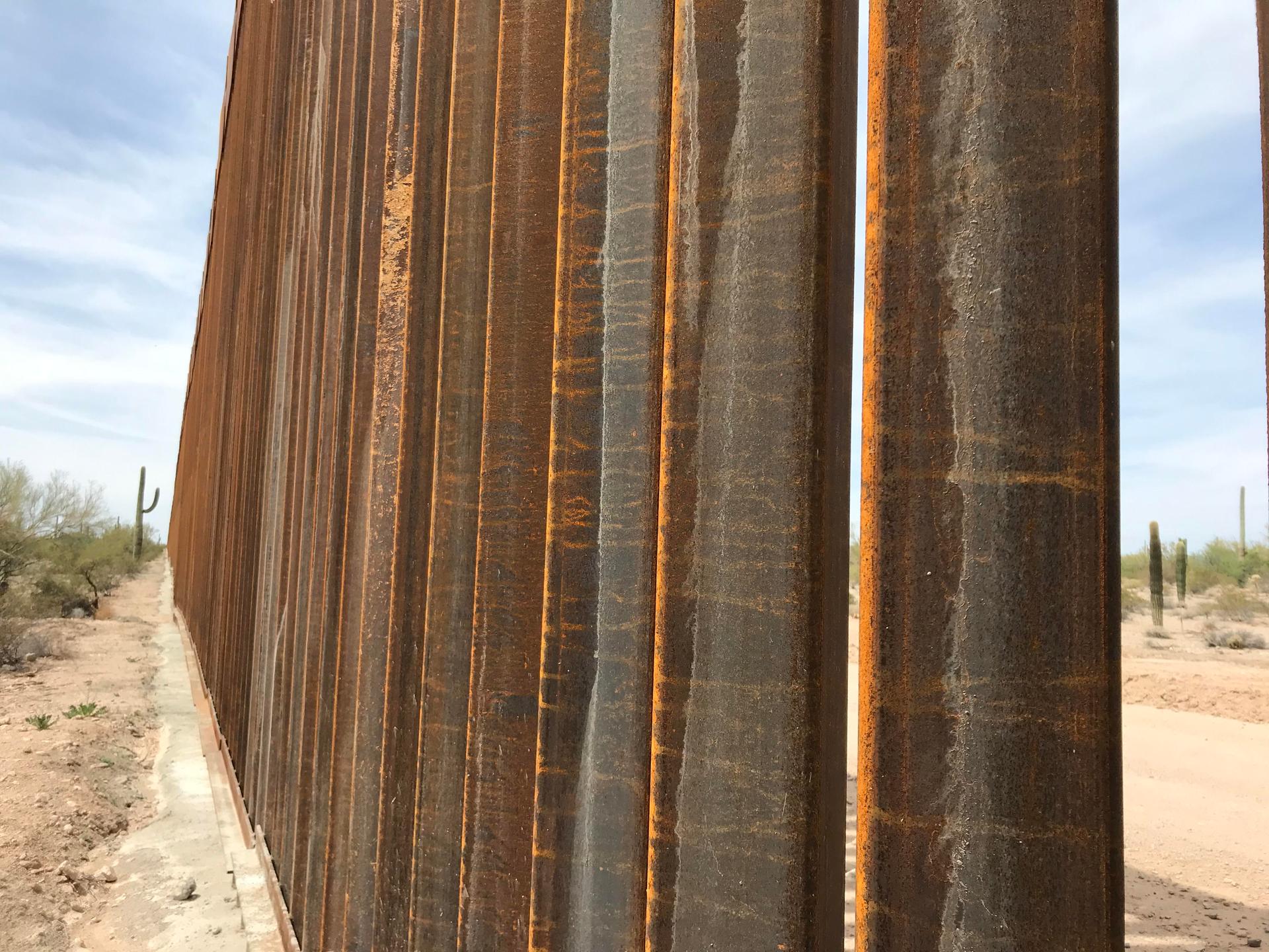 A view from the Mexican side of a 30-foot border wall, peeking through the slabs to show the US side of the border.