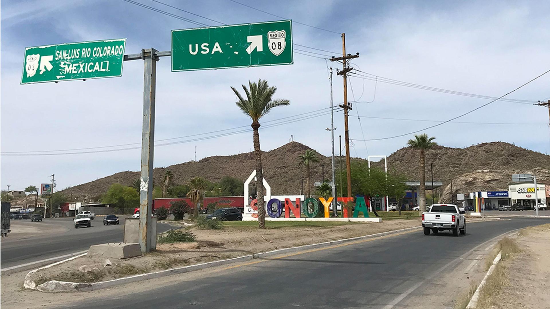 Street shot showing sign pointing to the USA, with a white truck driving down road, and colorful letters spelling Sonoyta, against backdrop of hills.