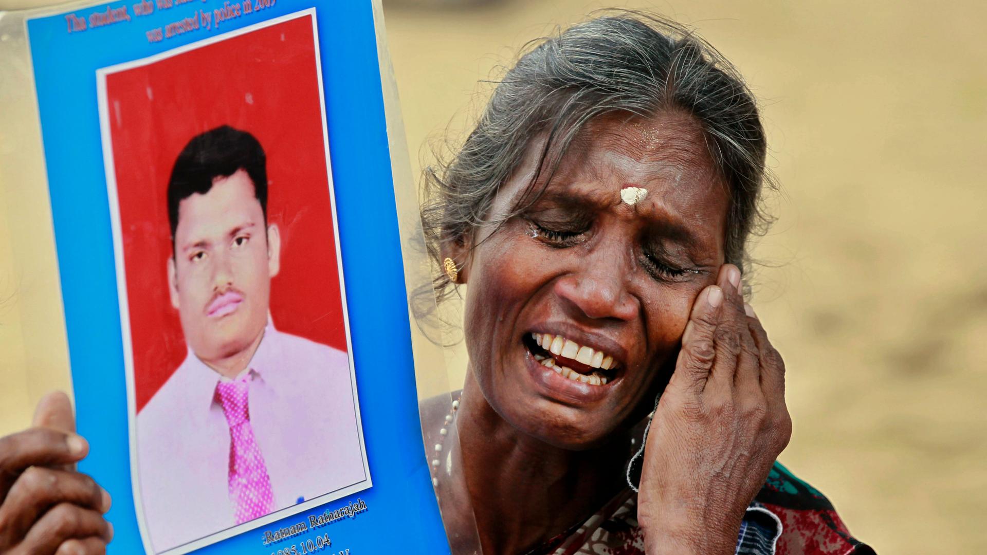 A woman weeps while holding a picture of her dead son, with a hand to her cheek, and eyes closed. 