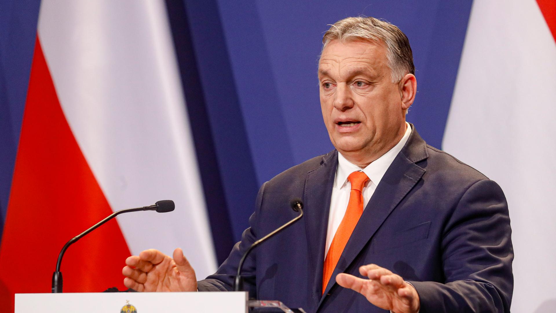 Hungarian Prime Minister Viktor Orban is shown wearing a blue suit and orange tie while speaking at a podium with two microphones.