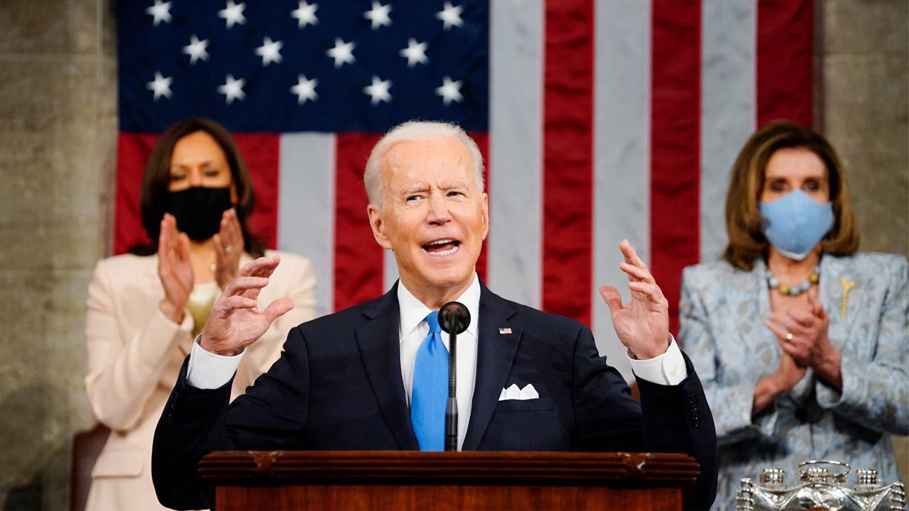 Biden lifts his hands at podium wearing a suit with Harris and Pelosi standing behind him, clapping. 