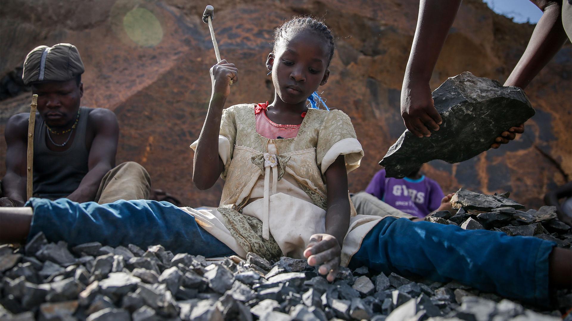 Young girl holds up hammer as she uses it to break rocks in front of her