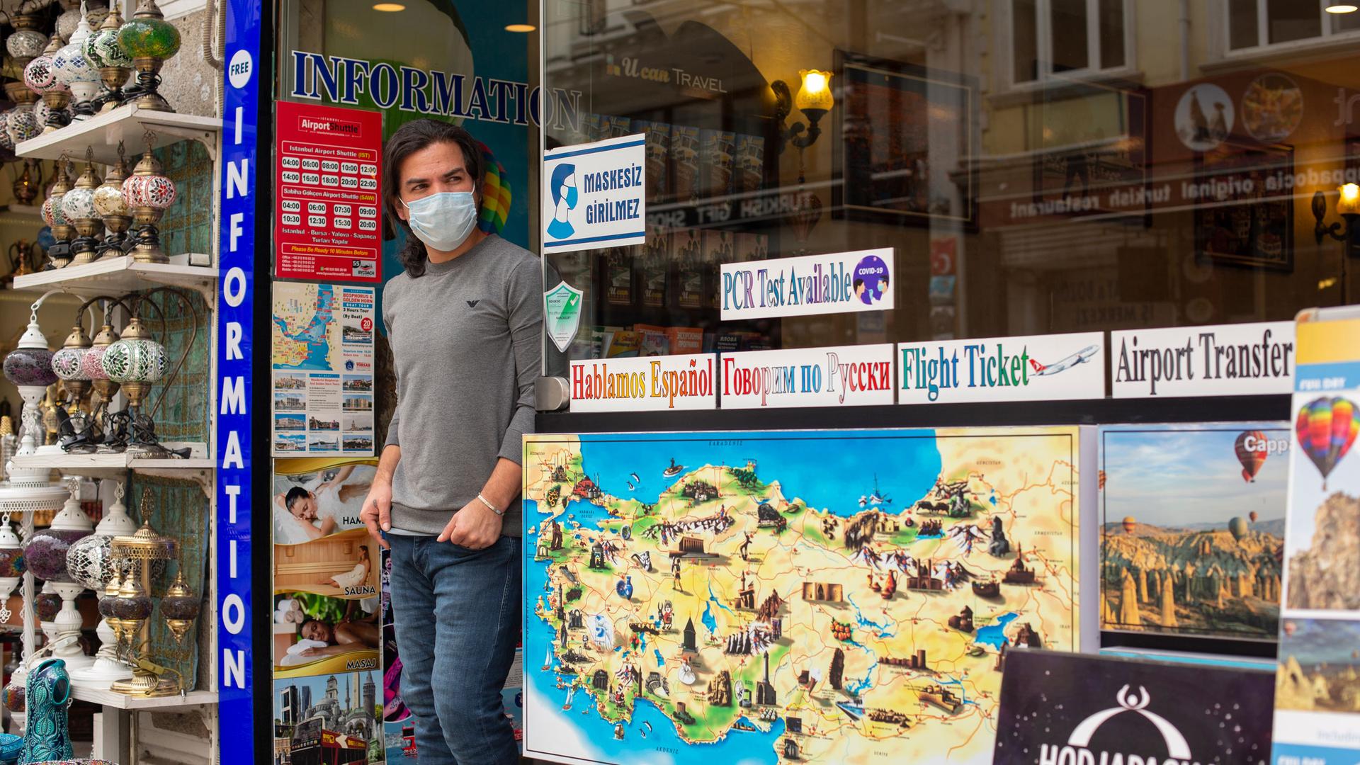 A man is shown wearing a face mask and standing in the doorway of a sightseeing store with maps of Istanbul on the windows.