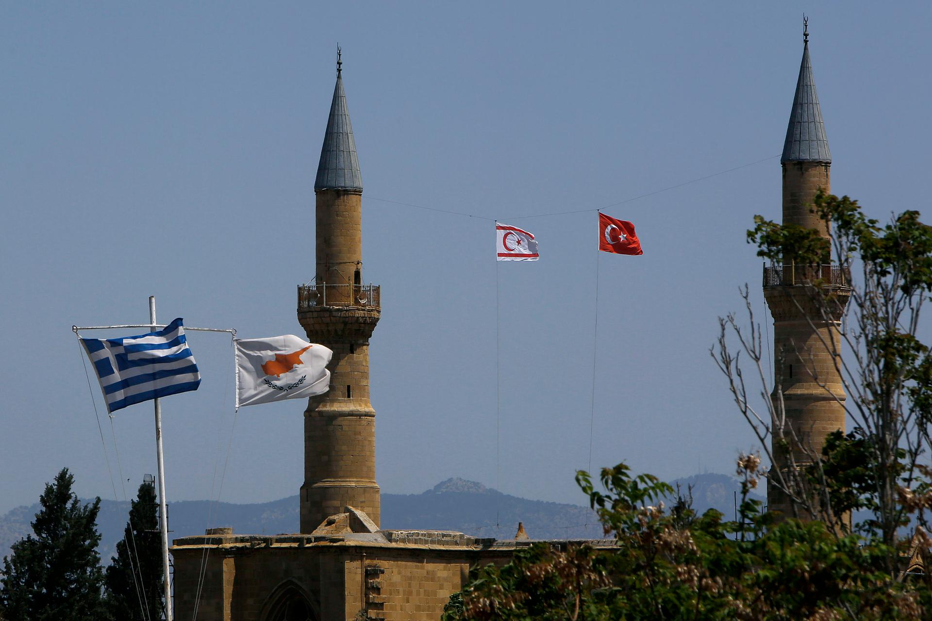 Greek and Cypriot flags flutter on poles on the left, as Turkish and Turkish Cypriot breakaway flags fly between minarets on the right
