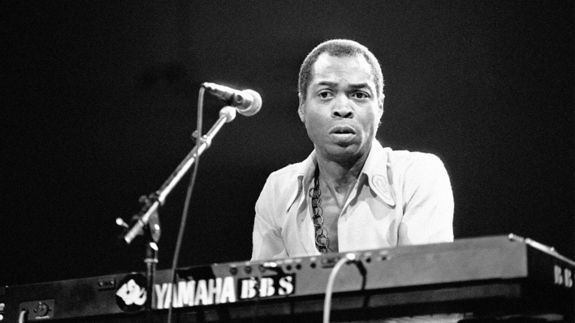 Fela Kuti is shown playing an electric keyboard and standing behind a microphone.
