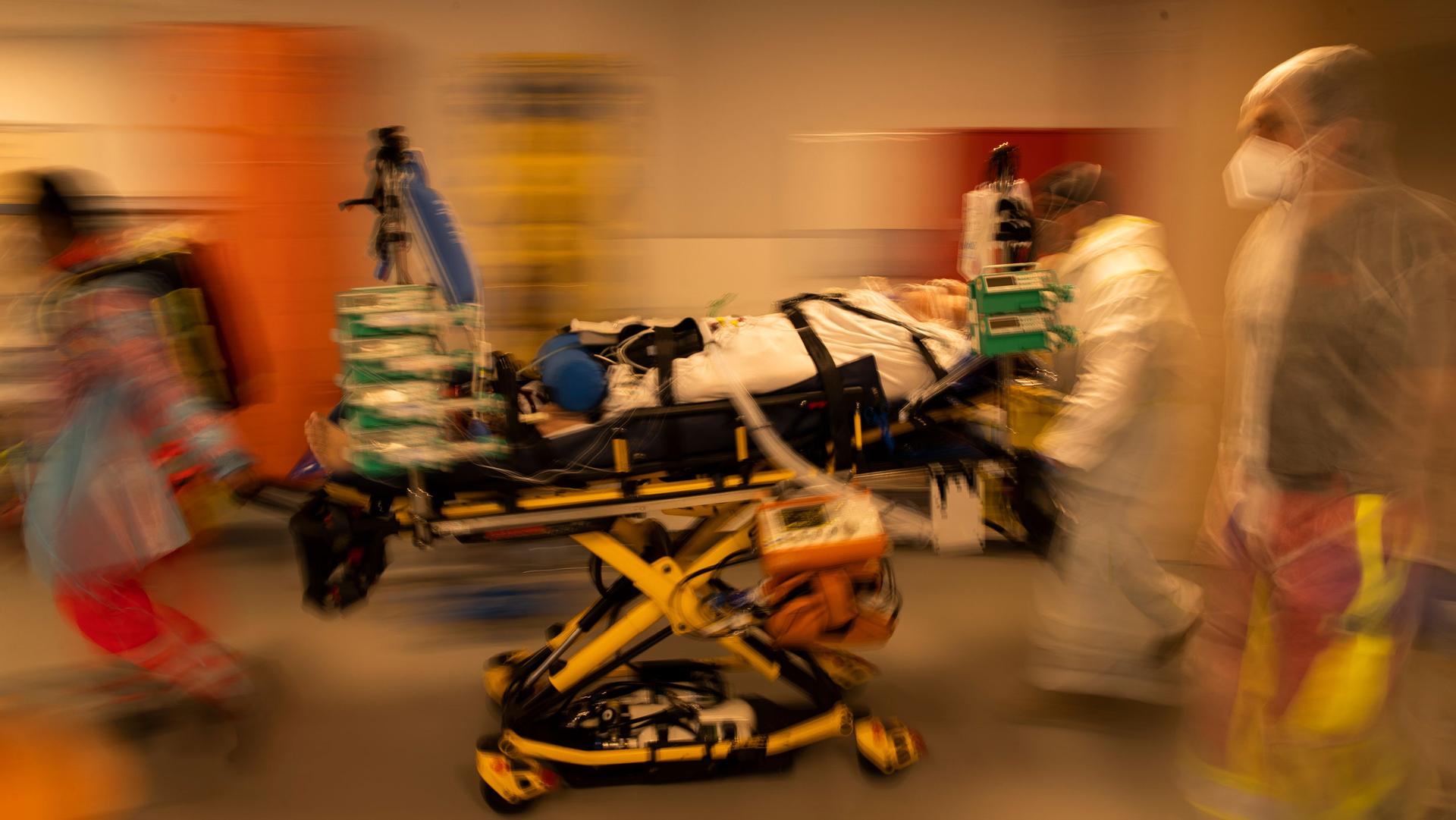 A COVID-19 patient is shown strapped to a yellow gurney with several medical staff moving along side in a blurred motion photograph.