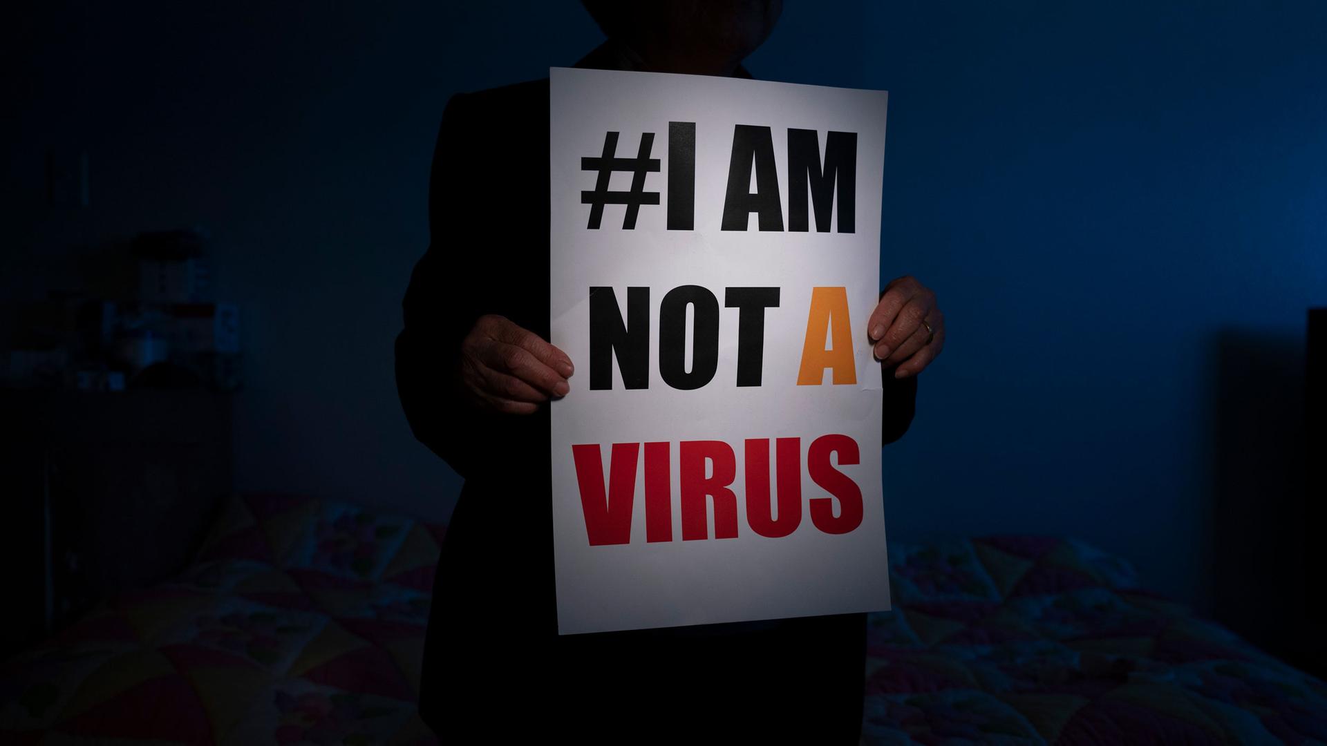A person is shown in shadow and holding a placard with "#I am not a virus" printed on it.