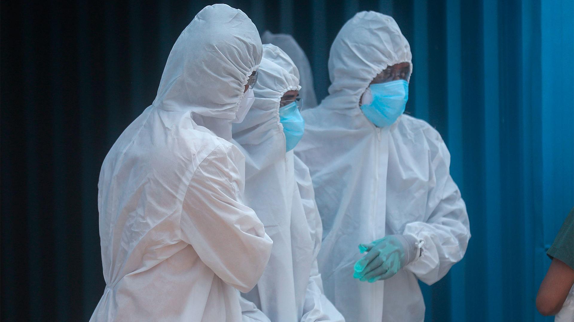 Three people wearing white biohazard suits look on to a dead relative out of frame