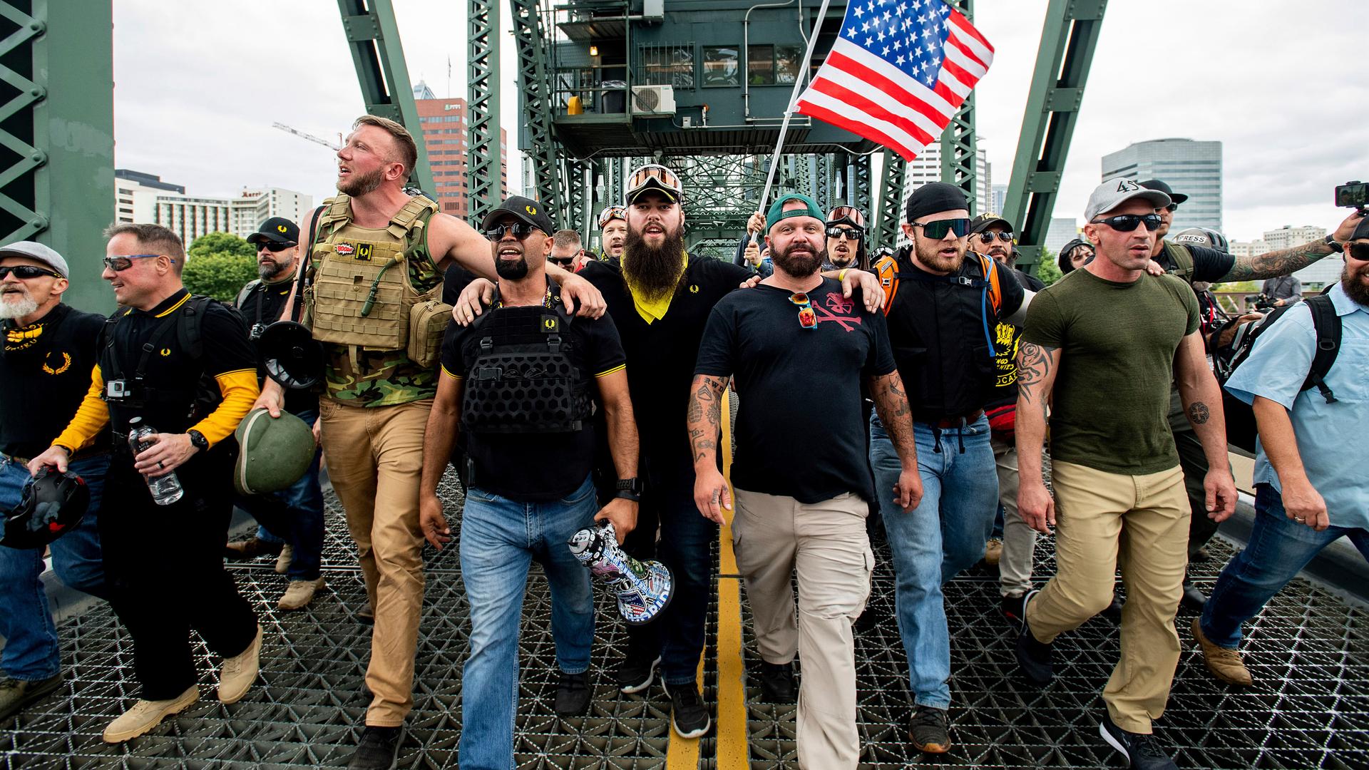 A group of men, mostly white, march with a US flag along a bridge. 