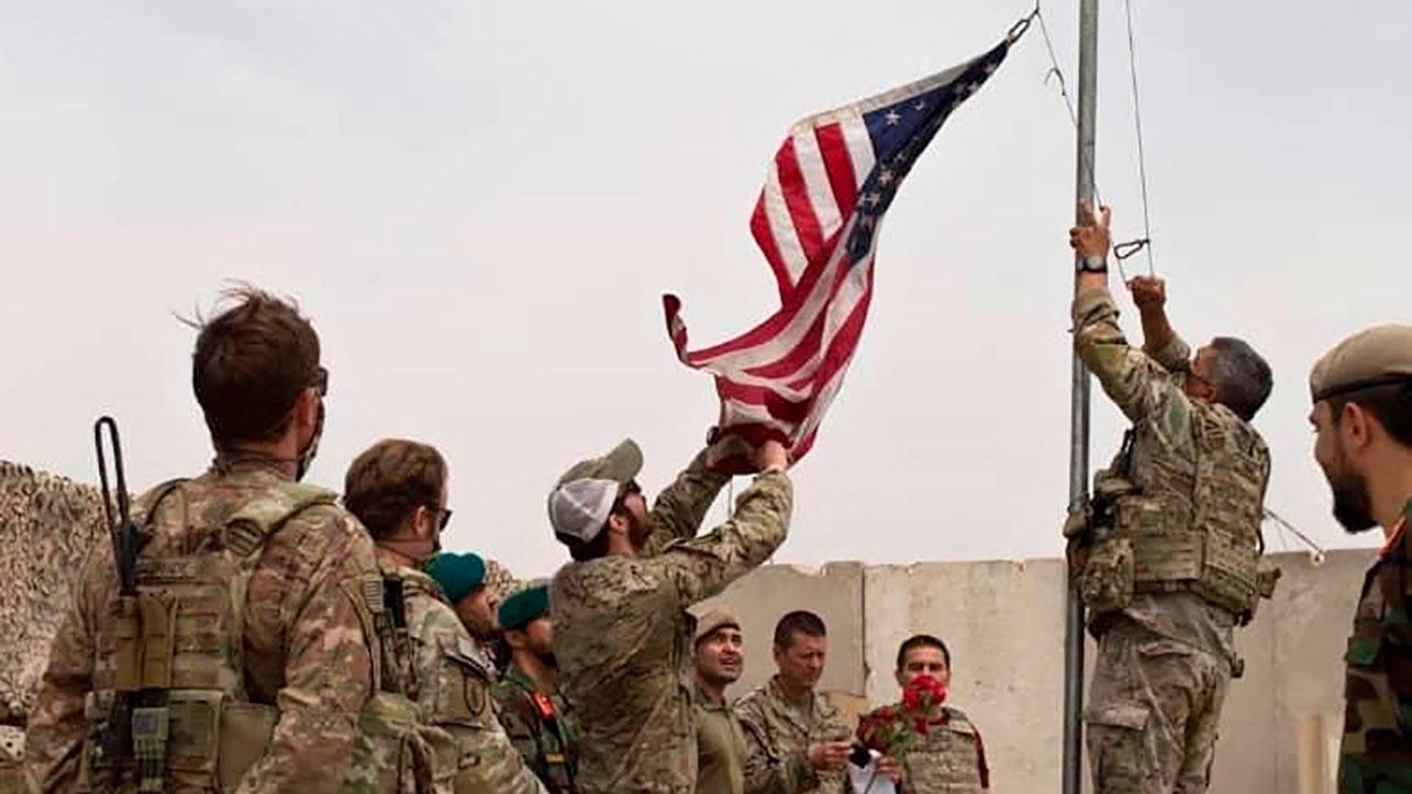 A small group of soliders are shown wearing military fatigues while one man is taking a US flag down off a flagpole
