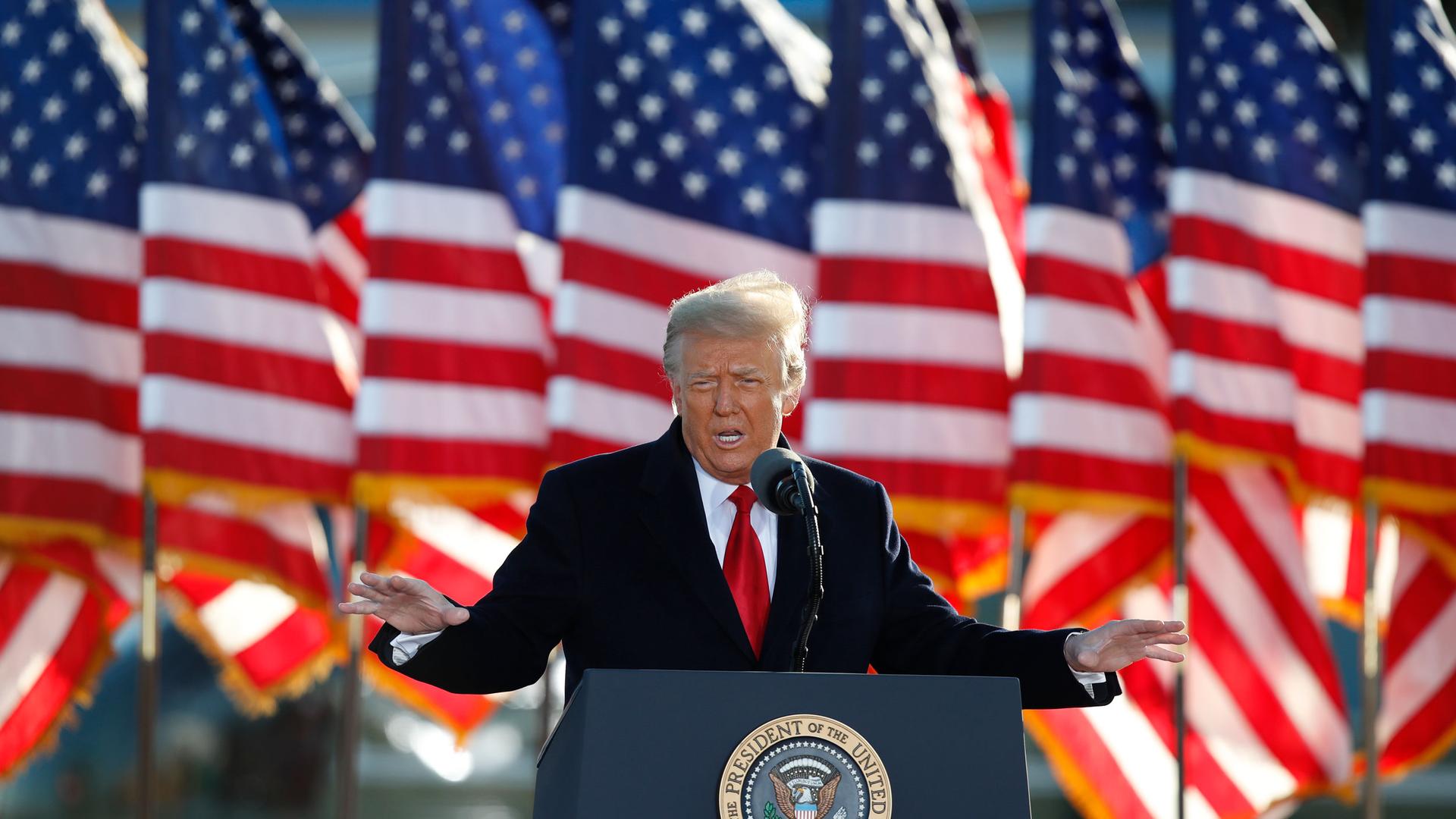 Donald Trump is shown wearing a dark suit overcoat and red tie with several US flags in the background.