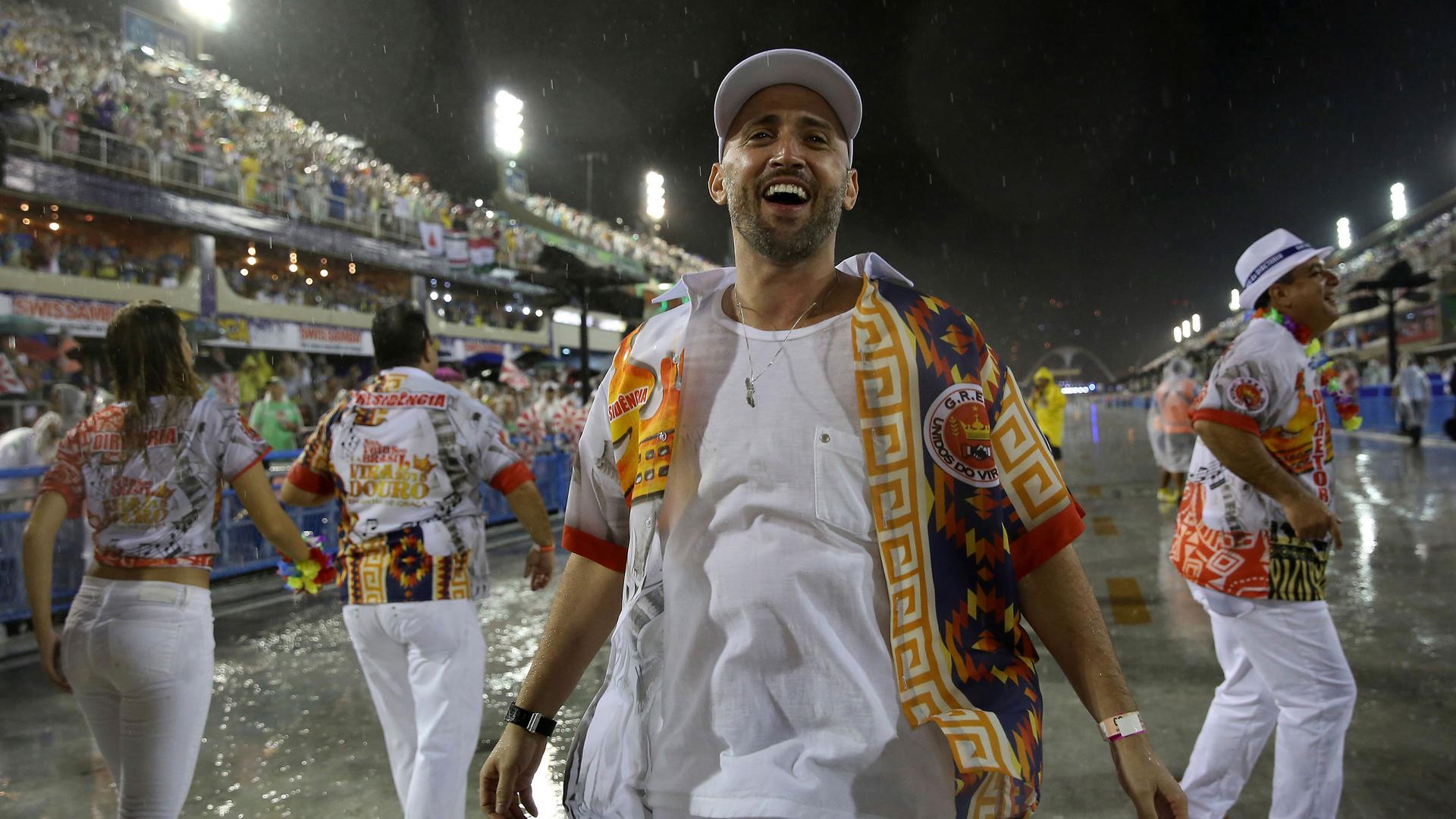 Comedian and actor Paulo Gustavo is shown wearing mostly white and brightly colored overshirt and smiling among other Carnival goers.