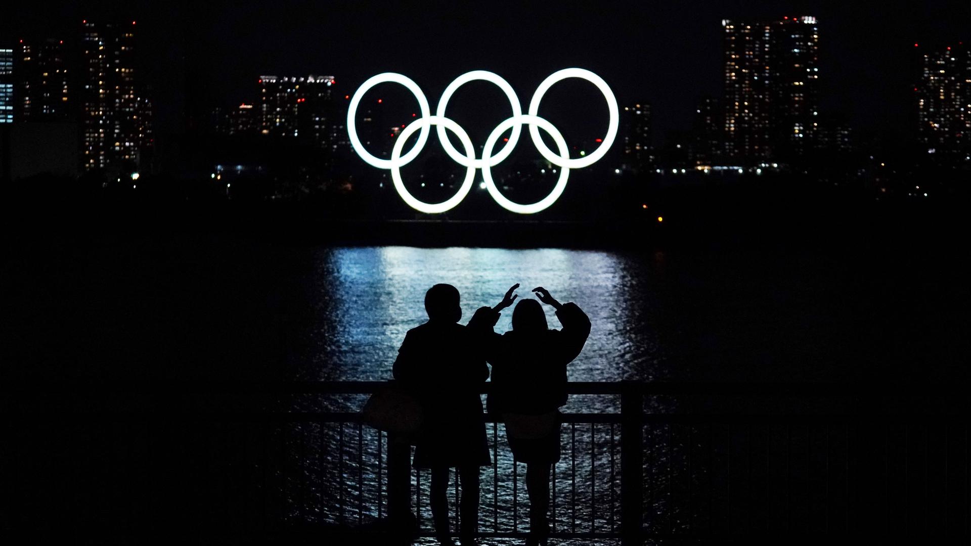 Two people are shown standing at a railing next to a body of water where the Olympic rings are in the destance illuminated in white.