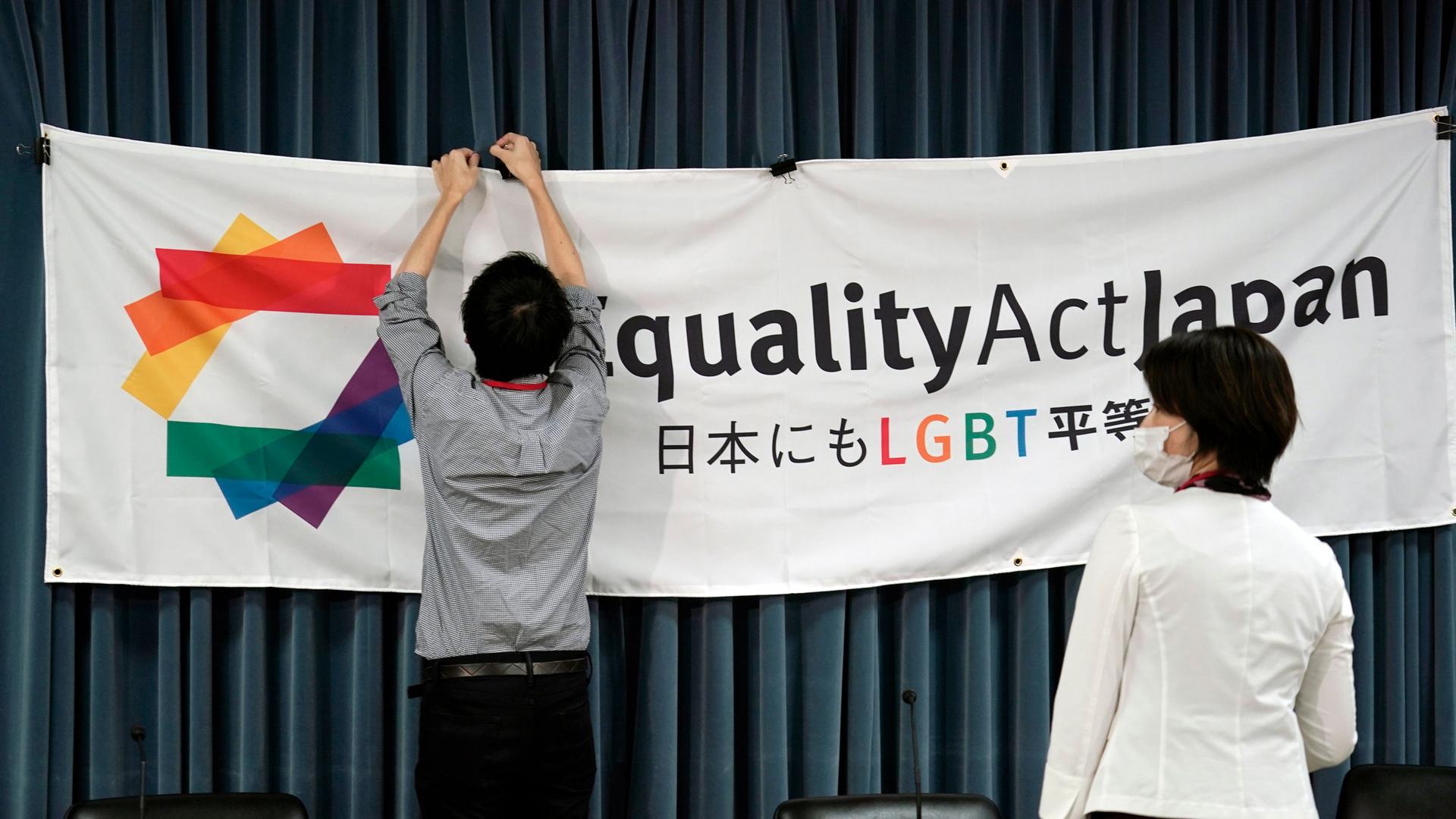 A person is shown clipping a large banner with the words, "Equality Act Japan" printed on it to a backdrop.