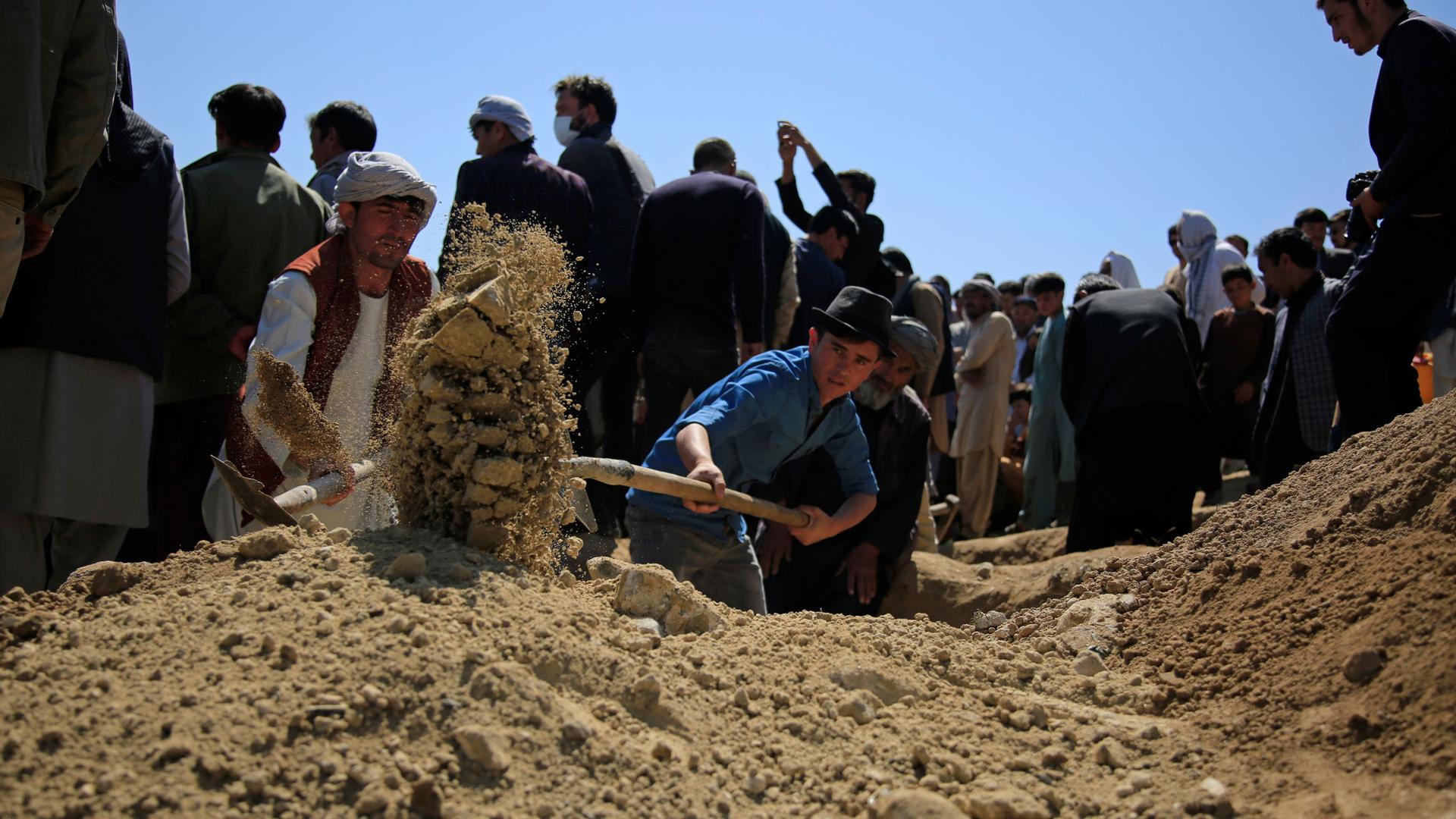 A large crowd of people are show at a cemetery as a man holds a shovel and is digging a burial site.