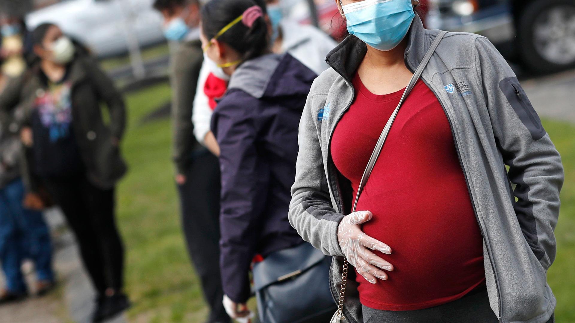 A pregnant person wearing a red shirt holds her belly, wearing a face mask and gloves. 