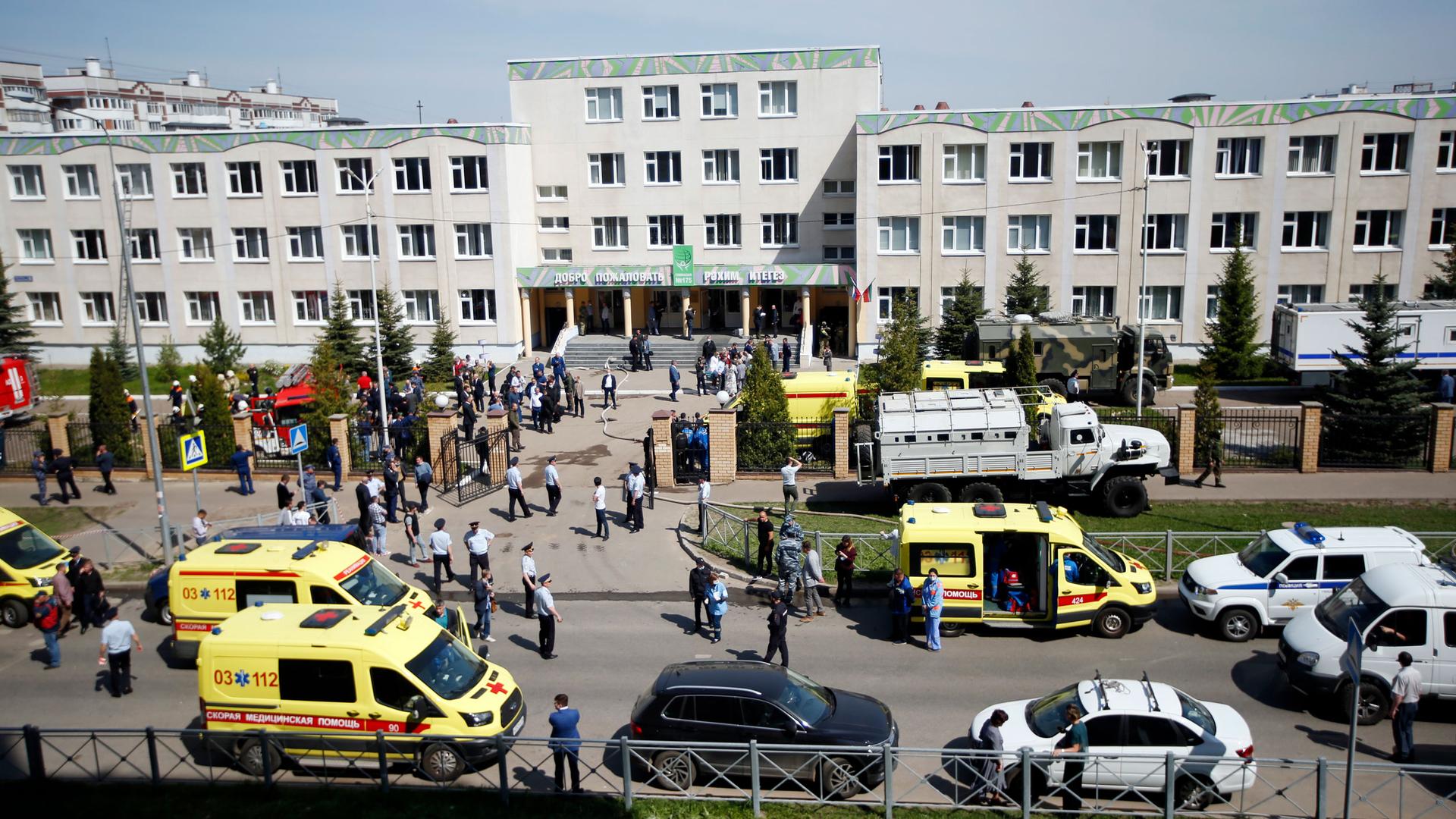 Several yellow ambulances and white police cars and are shown in the street outside of a three storey school building.