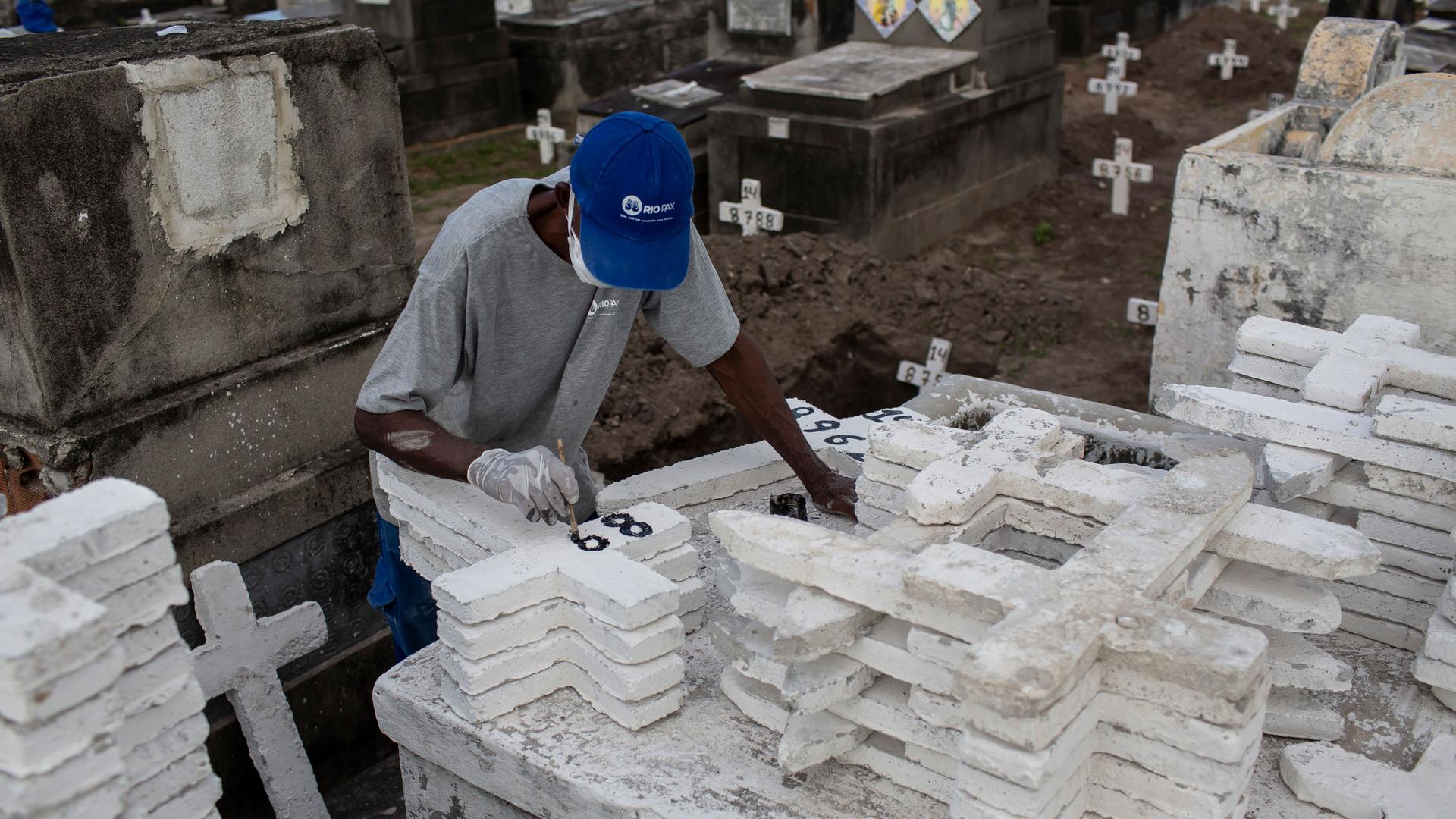 A man is shown wearing a blue hat and standing in front of dozens of white crosses while painting numbers on the crosses.