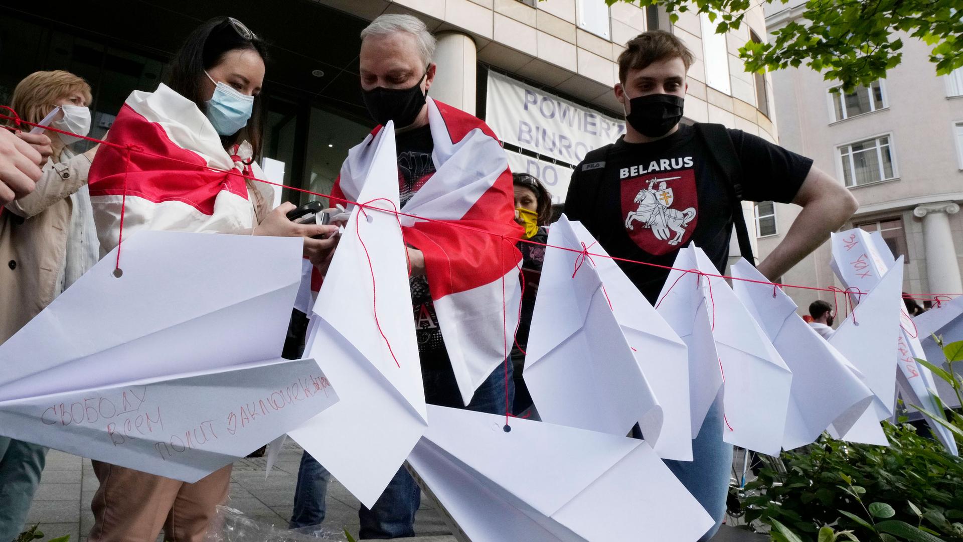 Several people are shown wearing protective face masks and standing behind a red string with several folded paper airplanes attached.