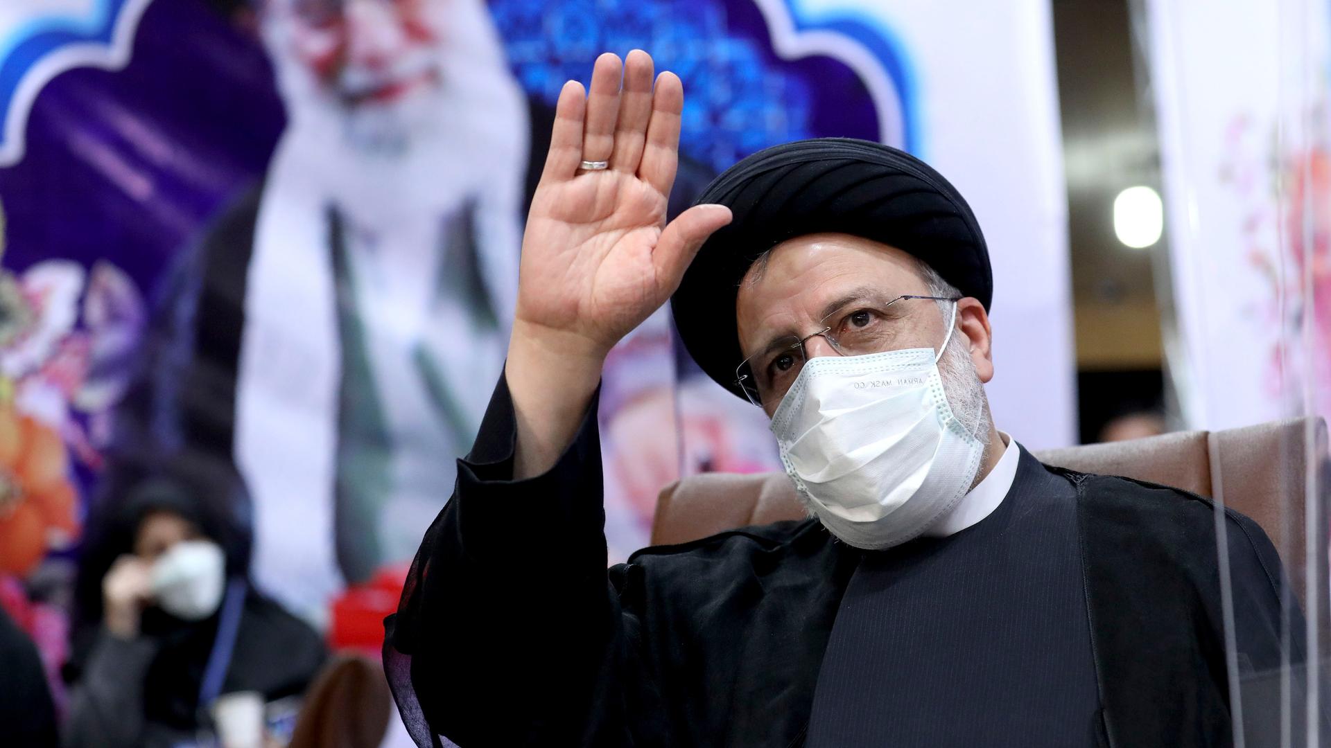 Iranian man in black robe and turban waves in front of a picture of Supreme Leader Ali Khamenei