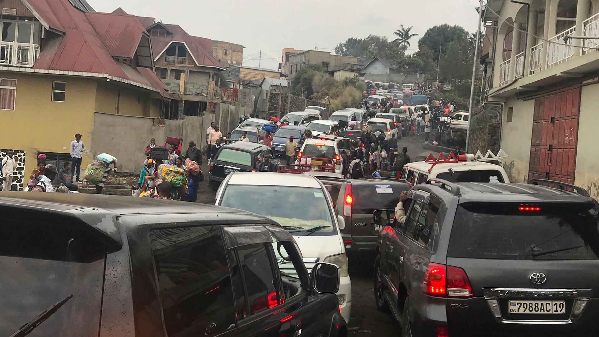 Dozens of cars are shown from behind packed into a road trying to drive out of Goma with houses on either side.