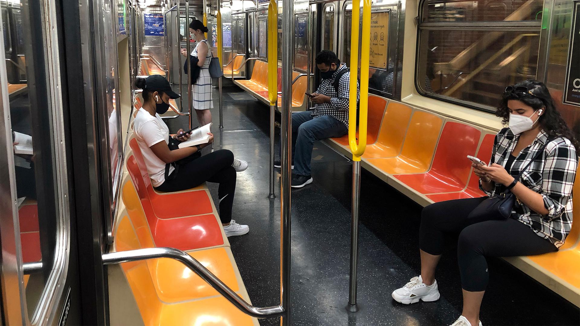 People wearing different types of black and white clothes sit and stand among orange and yellow seats on the New York subway