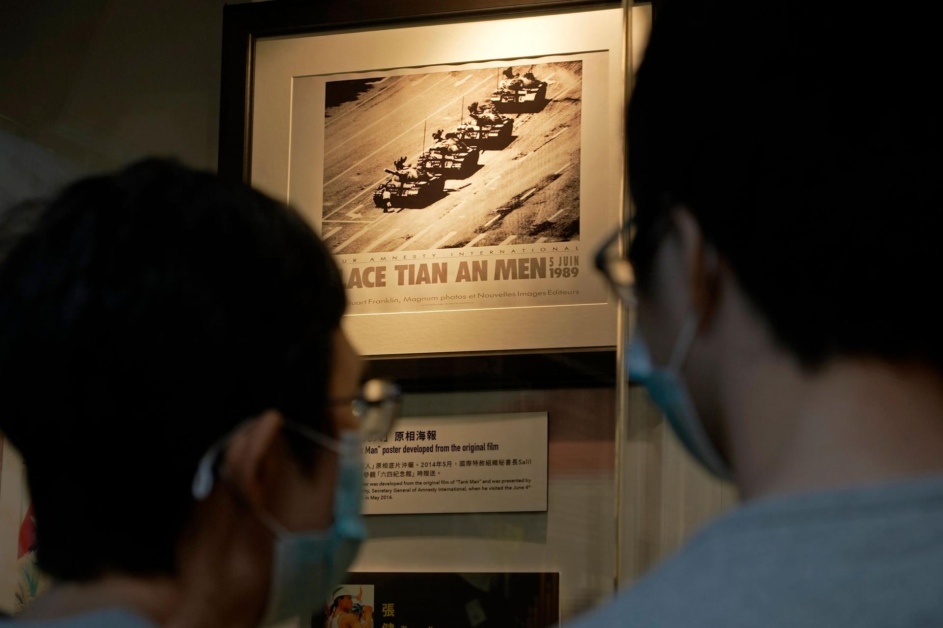 Two people are shown wearing protective face masks and looking at a photograph of a man blocking a row of four tanks.